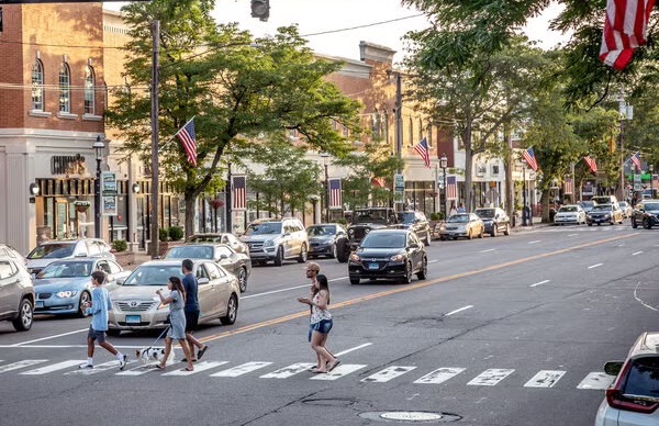 People crossing a downtown road past parked cars and shops
